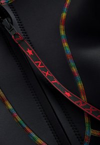 Black wetsuit material with multicolored zigzag stitching along seams. Features a black zipper with a red tag marked "NXT" and stars.