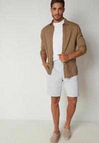 Camicia button-up marrone chiaro sopra una t-shirt bianca, abbinata a pantaloni corti grigi chiari e scarpe slip-on beige. Design semplice con una vestibilità rilassata.