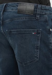Tmavomodré denimové džínsy so zadnou kapsou a malou vlajkou Tommy Hilfiger, čierna kožená štítok značky na opasku.