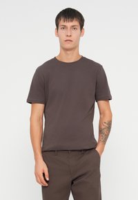 Kortærmet T-shirt med rund hals i mørkebrun bomuld, der har en glat overflade. Båret med mørkebrune bukser og en synlig arm tatovering.