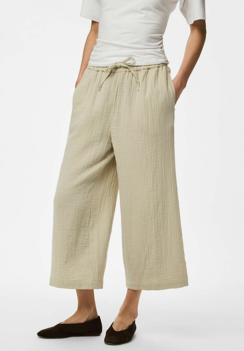 Femme portant un pantalon beige large et court avec taille à cordon et chaussures plates noires, mains dans les poches, debout devant un fond blanc.