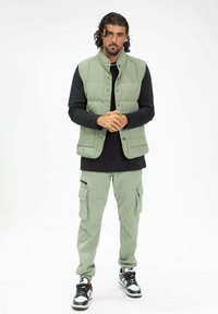 Gilet rembourré vert clair sur une chemise à manches longues noire, associé à un pantalon cargo assorti. Des baskets noires et blanches complètent la tenue.