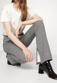 Mujer en cuclillas vistiendo pantalones de patrón jaquard en blanco y negro con aberturas laterales, camiseta blanca de manga corta y botas negras brillantes hasta el tobillo.