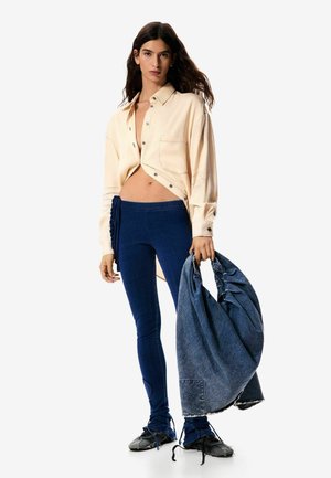 Mujer con camisa beige abierta, leggins azules, zapatos negros, sosteniendo una chaqueta de mezclilla arrugada grande contra un fondo blanco.