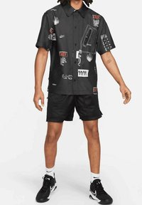 Chemise noire à manches courtes avec divers imprimés graphiques. Portée avec un short en mesh noir et des chaussures de sport noires, avec des accents blancs de la marque Nike.
