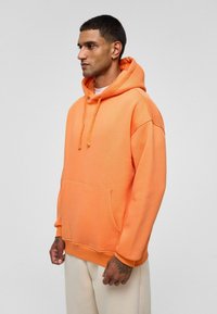 Oranje pullover hoodie van katoen, met een ruime voorzak, capuchon met koord en ribgebreide manchetten, gedragen met beige broek.