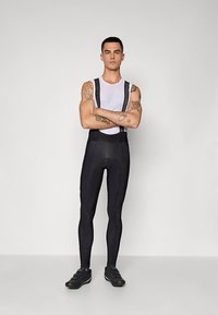 Homem de braços cruzados, vestindo calças de bib de ciclismo pretas, uma parte de cima de malha sem mangas branca, sapatos de ciclismo pretos e tatuagens visíveis nos braços.