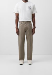 Bílý tričko s černou grafikou na levé hrudi, zkombinované se světle hnědými kalhotami s širokými nohavicemi a bílými teniskami. Jednoduchý a ležérní design.