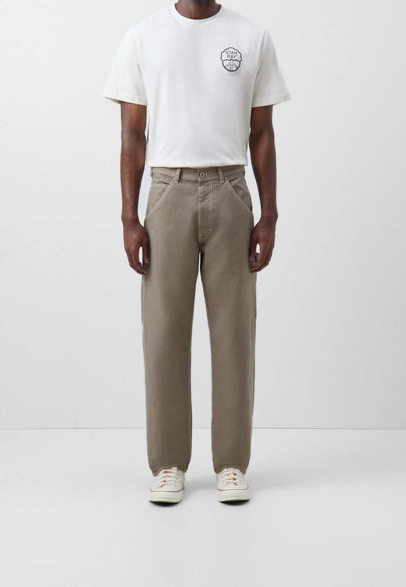 Bílý tričko s černou grafikou na levé hrudi, zkombinované se světle hnědými kalhotami s širokými nohavicemi a bílými teniskami. Jednoduchý a ležérní design.