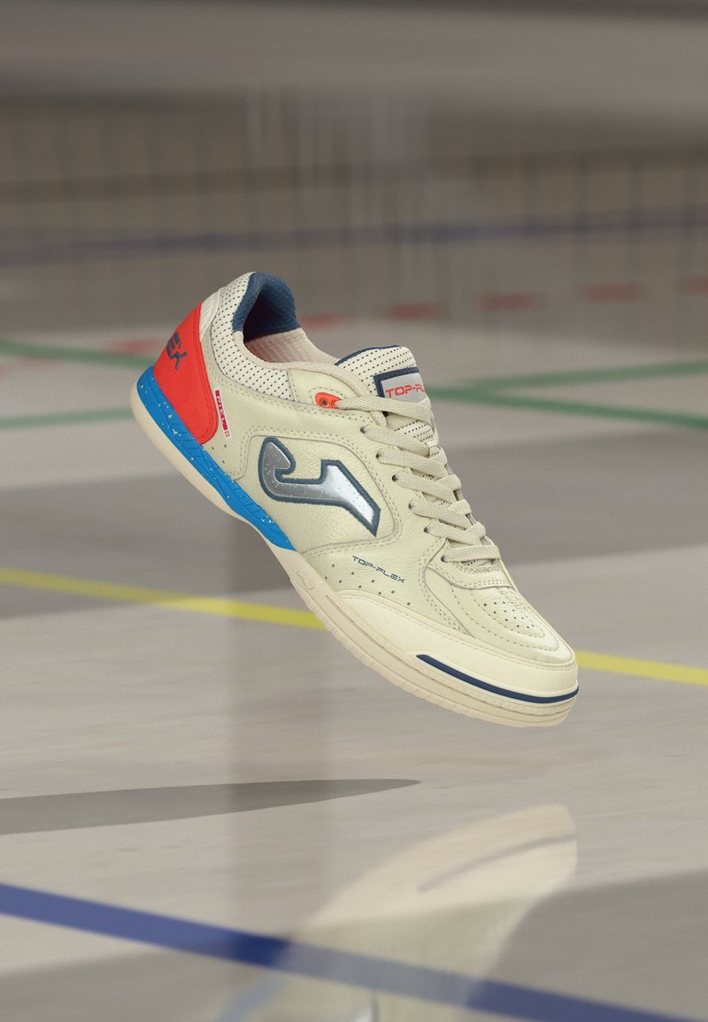 Λευκό παπούτσι για ποδόσφαιρο σε εσωτερικό χώρο με μπλε και κόκκινες λεπτομέρειες και κορδόνια, να αιωρείται πάνω από το πάτωμα ενός γυμναστηρίου με χρωματιστές γραμμές γηπέδου.