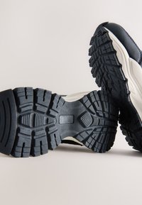 Hvide og marineblå fritidssko med en tekstureret sort gummisål med dybe mønstre, lagdelt design og åndbart netoverdel.