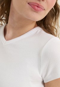 Bílé proužkované tričko s výstřihem do V a krátkými rukávy. Látka vykazuje jemné vertikální pruhy, což vytváří texturovaný vzhled.