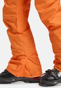Oranžové nepresakujúce snehové nohavice zakrývajúce čierne lyžiarske topánky s nastaviteľnými prackami, navrhnuté na zimné športy a ochranu pred chladným počasím.