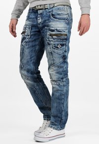 Jeans in denim blu sbiadito con numerose tasche con zip, vestibilità slim e cuciture decorative lungo le cuciture. Abbinati a sneakers bianche.