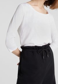 Witte lange mouwen top met een bootnek, gecombineerd met zwarte broek met elastische taille en trekkoord, voorzien van zijzakken. Soepele textuur.