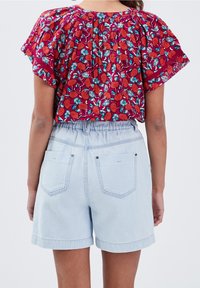 Donna che indossa pantaloni denim a vita alta di colore azzurro chiaro e una maglietta rossa floreale a maniche corte e svasata, vista di spalle contro uno sfondo neutro.
