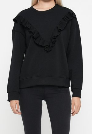 Kobieta ubrana w czarną, długorękawową bluzę z falbankami ułożonymi w kształt litery V na przodzie oraz czarne dopasowane spodnie.