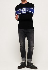 Uomo che indossa un maglione nero con strisce blu e bianche e la scritta "Superdry", jeans scuri arrotolati e stivali neri con lacci.