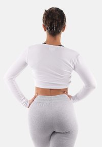 Weißes langärmliges Crop-Top mit gerippter Textur, kombiniert mit hellgrauen hochtaillierten Leggings. Der Hintergrund ist rein weiß.
