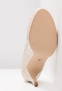 Chaussure à talon beige inclinée avec une semelle lisse, un bout pointu et un petit talon sur un fond blanc à angle.