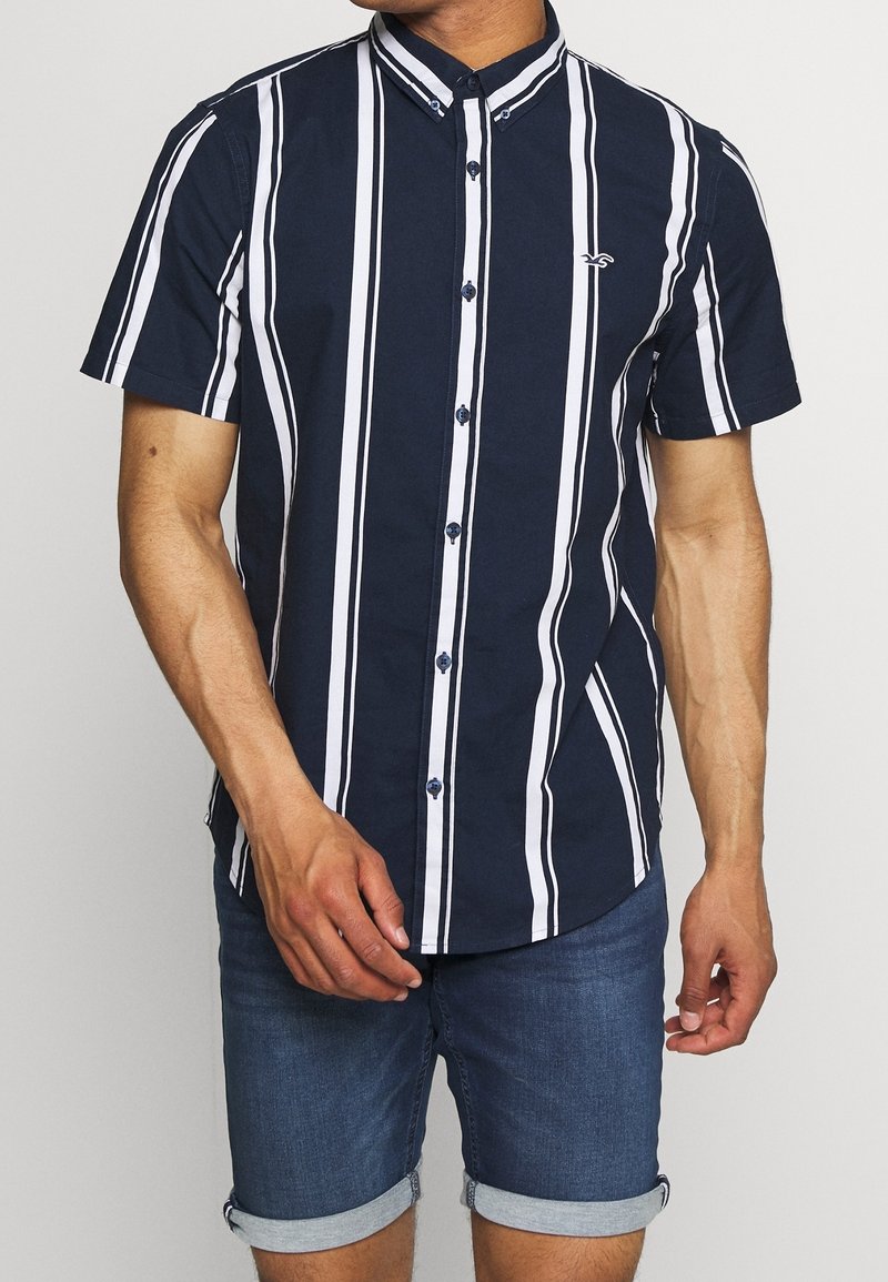 Marinblå kortärmad knapp-up skjorta med vita vertikala ränder, en knapp-ned krage och en liten logotyp på bröstet. Bomullsblandning.