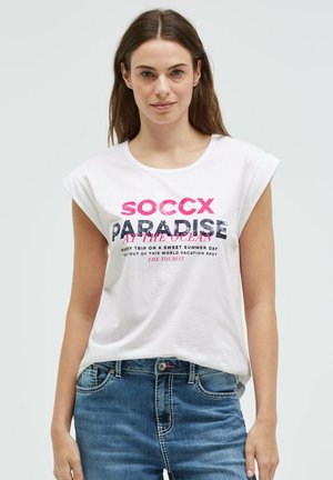 Young woman with long hair wearing a white sleeveless top with "SOCCX PARADISE AT THE OCEAN" text and blue jeans, standing against a plain background.
