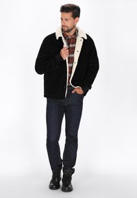Sort fløjlsjakke med hvid pelskrave, forreste knapper og ternet skjorte underneath. Mørke jeans og sorte læderstøvler fuldender outfittet.