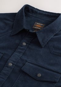 Marineblå kordfløjls skjorte med knaplukning ved kraven, to brystlommer og metal knapper. Har et struktureret stof og en mærket indvendig label.