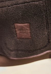 Braunes Fleecegewebe mit einer strukturierten Oberfläche, versehen mit einem Lederpatch mit der Aufschrift "NEXT". Der untere Rand hat eine glatte, kontrastierende braune Umrandung.