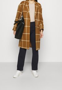 Brun rutigt ullmantel med en avslappnad passform, tillsammans med mörkblå byxor och vita sneakers. En svart väska med handtag fulländar looken.