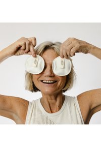 Dischetti di cotone bianchi con una piccola etichetta sono tenuti davanti al volto sorridente di una donna. Indossa un top bianco senza maniche e dalla trama lavorata.