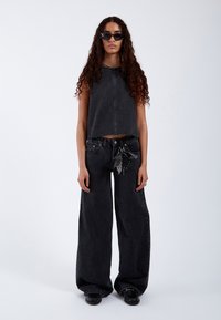 Vrouw met lang krullend haar, gekleed in een zwart mouwloos topje, zwarte wijde jeans met sleutelhangeraccessoire, zwarte schoenen en donkere zonnebril.