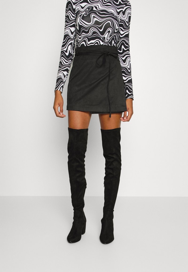 Černá semišová zavinovací sukně s páskem v pase, doplněná černými texturovanými kozačkami nad kolena. Dlouhý rukáv topu zdobí vlnitý černo-bílý vzor.