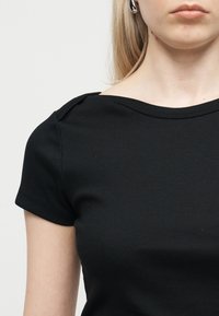 Νεαρή γυναίκα που φοράει μαύρο κοντομάνικο ριμπ κοντό τοπ με ελαφρώς φαρδύ ντεκολτέ και ασημένιο κρίκο στο αυτί.