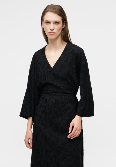 Robe noire avec un décolleté en V et des manches longues évasées. Fabriquée en tissu texturé présentant un motif floral subtil. Cintrée à la taille.