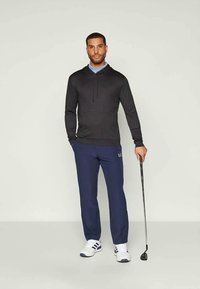 Moški v črni kapuci, modrih hlačah in belih teniskah stoji in drži golf palico pred enotno ozadjem.