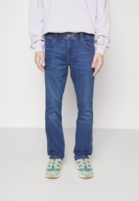 Blå denimjeans med slim fit, fem fickor och nitar, tillsammans med gröna sneakers med ett mönstrat design.