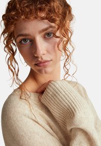 Jasnobeżowy sweter z dzianiny z prążkowanymi mankietami i okrągłym dekoltem, charakteryzujący się miękką teksturą i lekko luźnym krojem, noszony z jedną ręką na ramieniu.