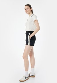Hvid polo skjorte med en sort krave, parret med sorte shorts med hvide sidelister. Modellen bærer hvide sokker og sneakers.