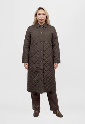 Une femme présente un long manteau marron foncé matelassé à losanges et boutonné, porté avec un pantalon en cuir et des bottines, sur un fond uni.