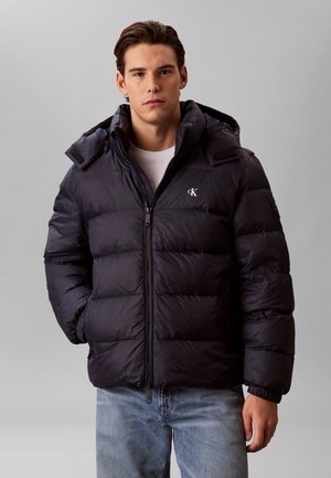 Jonge man die een zwarte pufferjas met capuchon, wit T-shirt en lichtblauwe jeans draagt, staande tegen een effen lichtgrijze achtergrond.