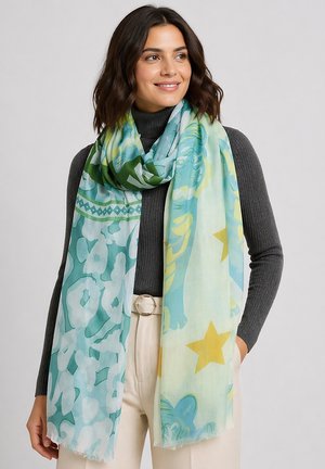 Frau mit welligem dunklem Haar, die einen dunkelgrauen Rollkragenpullover und hellbeige Hose trägt, lächelnd, mit einem großen pastellfarbenen Schal mit abstrakten blauen und gelben Sternen.