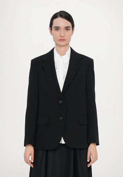 Sort blazer med en struktureret design, to store knapper og to forlommer. Båret over en hvid skjorte, der har en glat stoftekstur.
