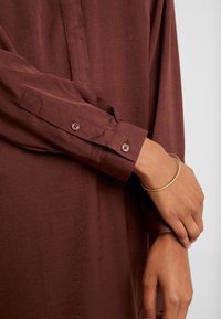 La main droite ajuste le poignet gauche portant un bracelet en or fin, les deux mains et avant-bras étant couverts par une chemise à manches longues marron à boutons.