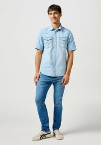 Camisa de mezclilla de manga corta en azul claro con dos bolsillos delanteros, costuras detalladas y un dobladillo curvado, combinada con jeans ajustados azules y zapatillas.