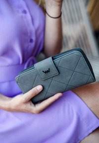 Gray wallet in textured material featuring a diamond pattern, a black logo on the clasp, and a zip closure, held in a hand against a purple dress.