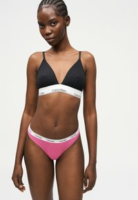 Femme portant un soutien-gorge triangle noir et un slip rose avec des bandes blanches portant le logo Calvin Klein sur un fond uni clair.