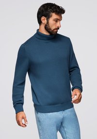 Tropfenblau gestrickter Rollkragenpullover mit gerippten Ärmelbündchen und Saum, aus strukturiertem Stoff und mit lässiger Passform, kombiniert mit hellblauen Jeans.