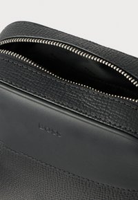 Μαύρη τσάντα από δέρμα με υφή, με κλείσιμο με φερμουάρ, που διαθέτει ασημένιες μεταλλικές λεπτομέρειες, λείες αποχρώσεις και διακριτική λεπτομέρεια με λογότυπο στο πλάι.