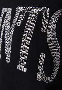 Sort stof med nærbillede af sølvhvidt broderi, der danner stiliserede store bogstaver i et fedt mønster.